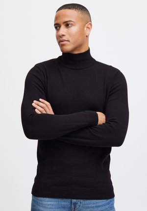 Suéter negro de cuello alto hecho de tela suave, con mangas largas y puños y dobladillo acanalados. Lleva puesto unos jeans de mezclilla azul claro.