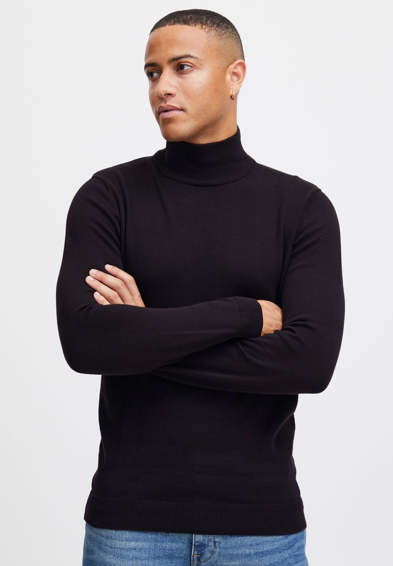Suéter negro de cuello alto hecho de tela suave, con mangas largas y puños y dobladillo acanalados. Lleva puesto unos jeans de mezclilla azul claro.