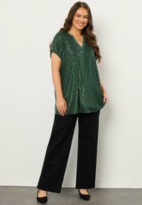 Tunika verde con paillettes, dal collo a V, maniche corte e una silhouette ampia, abbinata a pantaloni neri a gamba larga e tacchi neri.