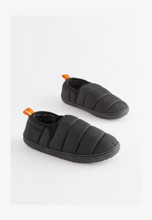 Mustat tikatut tohvelit, joissa on pehmeä, teksturoitu vuori. Varustettu mustalla kumipohjalla ja oranssilla vetokahvalla kantapäällä helpottamaan pukeutumista.