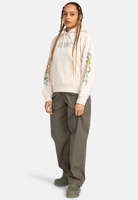 Felpa leggera color beige chiaro con ricami floreali sulle maniche, tasca a marsupio, pantaloni baggy verdi oliva abbinati e calzature verdi.