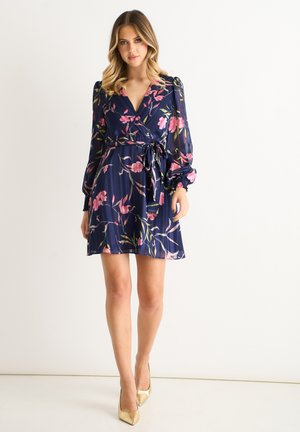 Woman wearing a navy blue floral wrap dress with long sheer sleeves and a waist tie, paired with gold pointed-toe high heels, standing against white background.