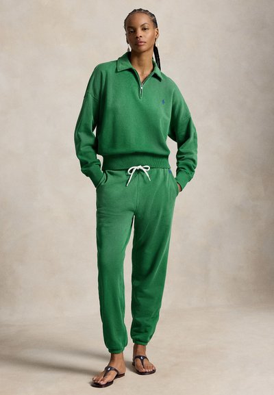 Kobieta z warkoczami, ubrana w zieloną bluzę z półzamkiem i spodnie dresowe z troczkami, stojąca z rękami w kieszeniach, mająca na sobie czarne sandały.