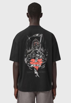 Personne de dos, portant un t-shirt noir avec un dessin de la Faucheuse tenant un cœur en sang transpercé par une dague et une bannière indiquant "L.D.A.".