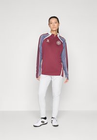 Maroone langærmet zip-up trøje med blåt stribede ærmer, hvide accenter og et rundt logo; parret med hvide træningsbukser og sorte sneakers.