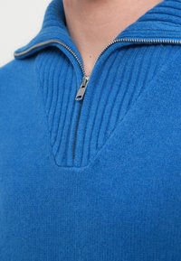 Pull bleu à fermeture éclair avec col côtelé et fini texturé. Le tissu semble doux et chaud, avec un motif en forme de V au niveau de l'encolure.