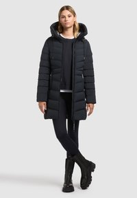 Zwart gewatteerde jas met capuchon, contrasterende zilveren voering, ritssluiting aan de voorkant en zijzakken. Gedragen over een donkerblauwe trui en zwarte legging.