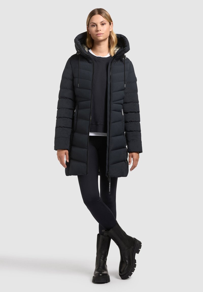 Haină puffer neagră cu glugă, cu un design până la genunchi, secțiuni matlasate și buzunare cu fermoar, purtată cu colanți negri și cizme masive.