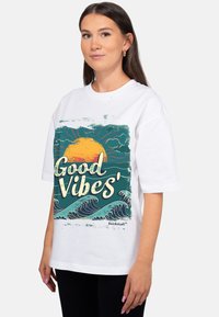 Wit T-shirt met een kleurrijke afbeelding van een zon, golven en de tekst "Good Vibes" in een speels lettertype. Losse pasvorm, korte mouwen.