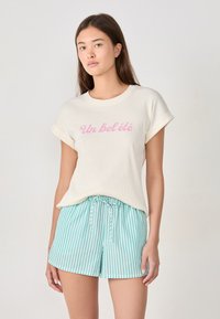 Camiseta de manga corta de canalé crema con mangas enrolladas que presenta el texto rosa "Un bel été", combinada con pantalones cortos a rayas verdes y blancas con cintura elástica y cordón.
