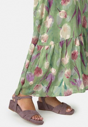 Nohy v růžově fialových semišových sandálech na klínovém podpatku s pásky kolem kotníků, částečně zakryté zelenými šaty s květinovým vzorem a volným střihem.