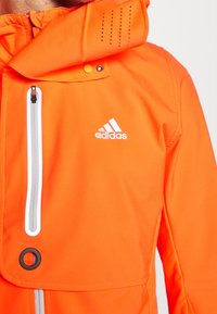 Giacca impermeabile arancione con zip completa, accenti bianchi a contrasto, cappuccio e dettagli perforati sulle spalle. Logo Adidas ben visibile.