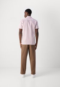 Camicia a maniche corte con righe verticali rosse e bianche, colletto classico e vestibilità rilassata, abbinata a pantaloni marroni e sneakers bianche.