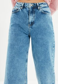 Vaaleansiniset korkeavyötäröiset farkut, joissa on napin ja vetoketjun sulkeminen, yhdistettynä vyötäröllä näkyvään vaaleanpunaisiin pitkälahkeisiin paitaan.