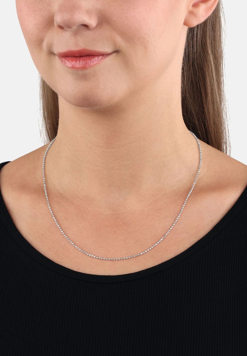 Donna che indossa una delicata collana di perline d'argento sopra un top nero, con il viso visibile nella parte inferiore, il collo e i lunghi capelli castani dietro l'orecchio.