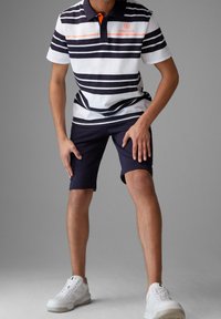 Polo rayé en blanc et bleu marine, avec des accents orange, associé à un short bleu marine et des baskets blanches. Accent sur un style décontracté sportif.