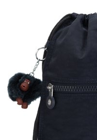 La mochila de tela azul marino cuenta con una parte superior tipo cordón, un bolsillo lateral con cremallera y un llavero de gorila negro y peludo sujeto con un clip de metal.