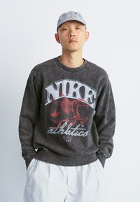 Nike sweatshirt i mørkegrå med stort rødt grafisk print, rund halsudskæring og ribbede manchetter. Sat sammen med hvide shorts.