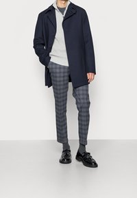 Manteau marine, pull à côtes gris clair et pantalon à carreaux gris. Chaussures habillées noires à lacets. Textures lisses, coupe ajustée.