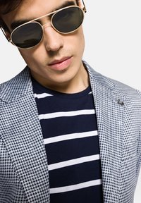 Szary żakiet w wzór houndstooth, granatowa i biała koszulka w paski oraz okrągłe okulary przeciwsłoneczne z ciemnymi soczewkami. Zdjęcie zbliżeniowe na rysy twarzy.