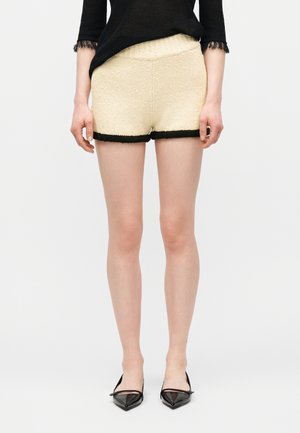 Frau trägt cremefarbene Strickshorts mit schwarzem Rand, kombiniert mit einem schwarzen Oberteil mit spitzenbesetzten Ärmeln und schwarzen spitzen Ballerinas.