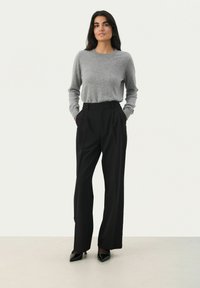 Grijze gebreide trui, zwarte geplooide broek, hoge hakken, model staat met handen in de zakken tegen een effen achtergrond.