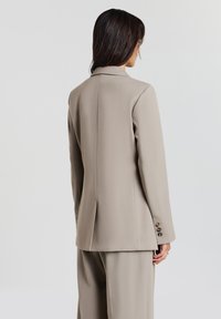 Donna con lunghe chiome scure, che indossa un blazer beige su misura e pantaloni abbinati, in piedi di spalle contro uno sfondo neutro.