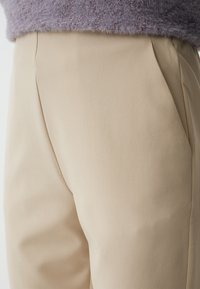 Pantaloni beige a vita alta con tessuto testurizzato, caratterizzati da pieghe frontali e un taglio pulito. Il materiale sembra morbido e leggermente strutturato.