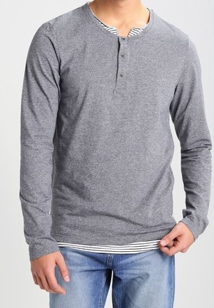 Un tee-shirt henley gris à manches longues avec un col rond, des boutons décoratifs et un ourlet à rayures superposé. Tissu doux avec une texture lisse.