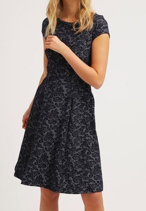 Blondynka w krótkiej, do kolan sukience z ciemnym kwiatowym wzorem i krótkim rękawem, stojąca na jednolitym tle.