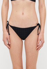 Bas de bikini noir avec un motif à carreaux texturé, des liens latéraux réglables et un design taille basse.