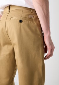 Khaki trousers with a smooth texture, featuring a rear pocket with a black button and a small brand tag on the waistband.
