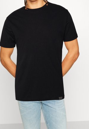 Homme portant un t-shirt noir uni à manches courtes et un jean bleu clair, debout les mains derrière le dos, sur un fond blanc uni.