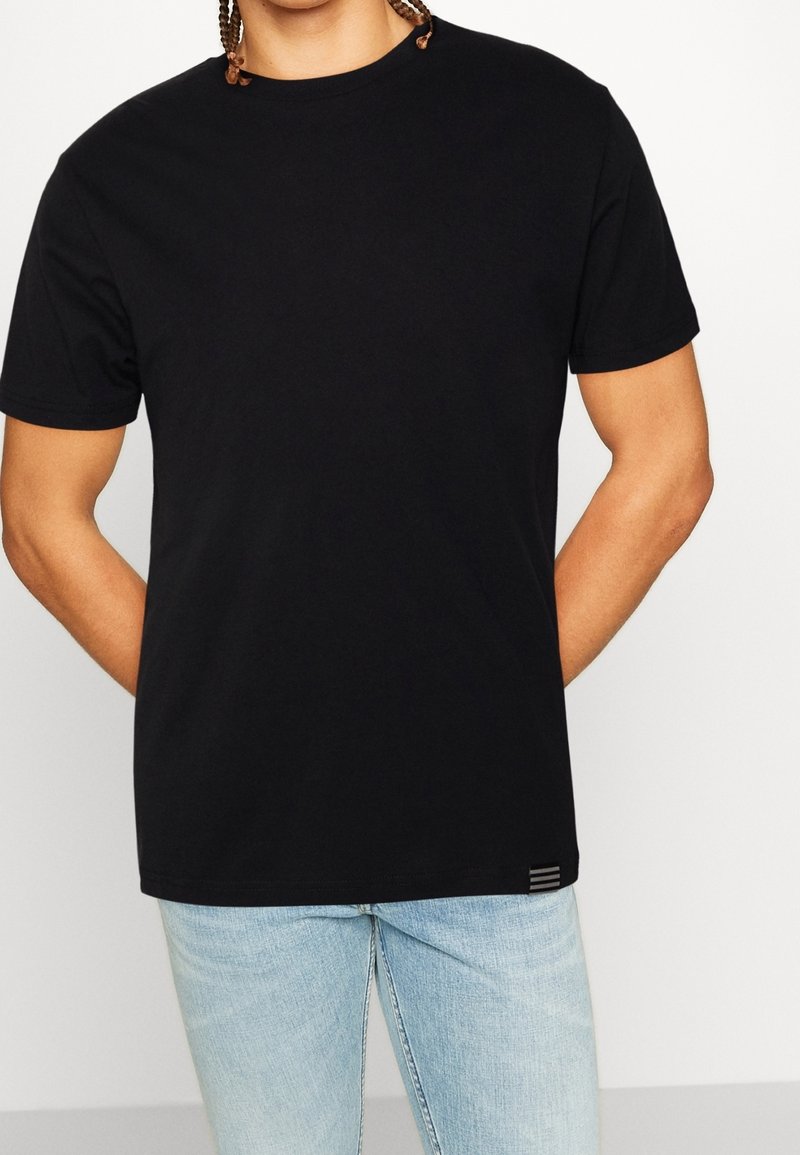 Homme portant un t-shirt noir uni à manches courtes et un jean bleu clair, debout les mains derrière le dos, sur un fond blanc uni.