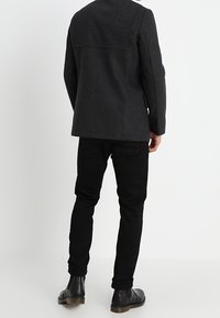 Zwarte wollen jasmix met een getailleerde pasvorm, voorzien van een rechte snit en minimale naden, gecombineerd met zwarte taps toelopende broek en enkellaarsjes.