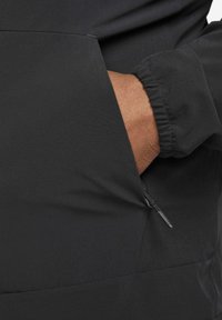 Zwarte jas met een zijzak met rits, elastische manchetten en een gladde, duurzame stoftextuur.