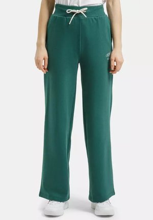 Osoba ubrana w ciemnozielone, szerokie spodnie dresowe z białym sznurkiem i białe sportowe buty, stoi na jednolitym tle.