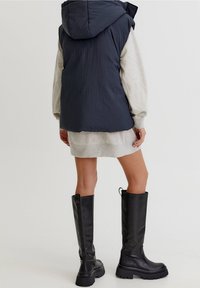Femme portant des bottes noires montantes, une robe sweat-shirt gris clair et un gilet sans manches matelassé bleu marine avec capuche, debout de dos.