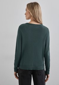 Zielony sweter z dzianiny, z długimi rękawami, okrągłym dekoltem oraz ściągaczami na mankietach i u dołu. Tekstura jest miękka i gładka, a fason dopasowany.