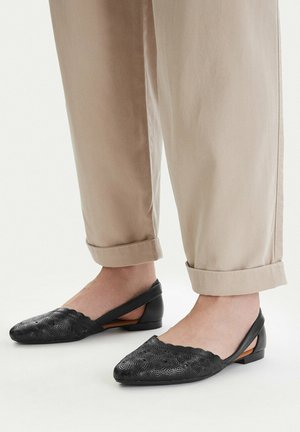 Stopy w beżowych spodniach z mankietami i czarnych skórzanych balerinach na niskim obcasie z kwiatowym wzorem, na białej podłodze.