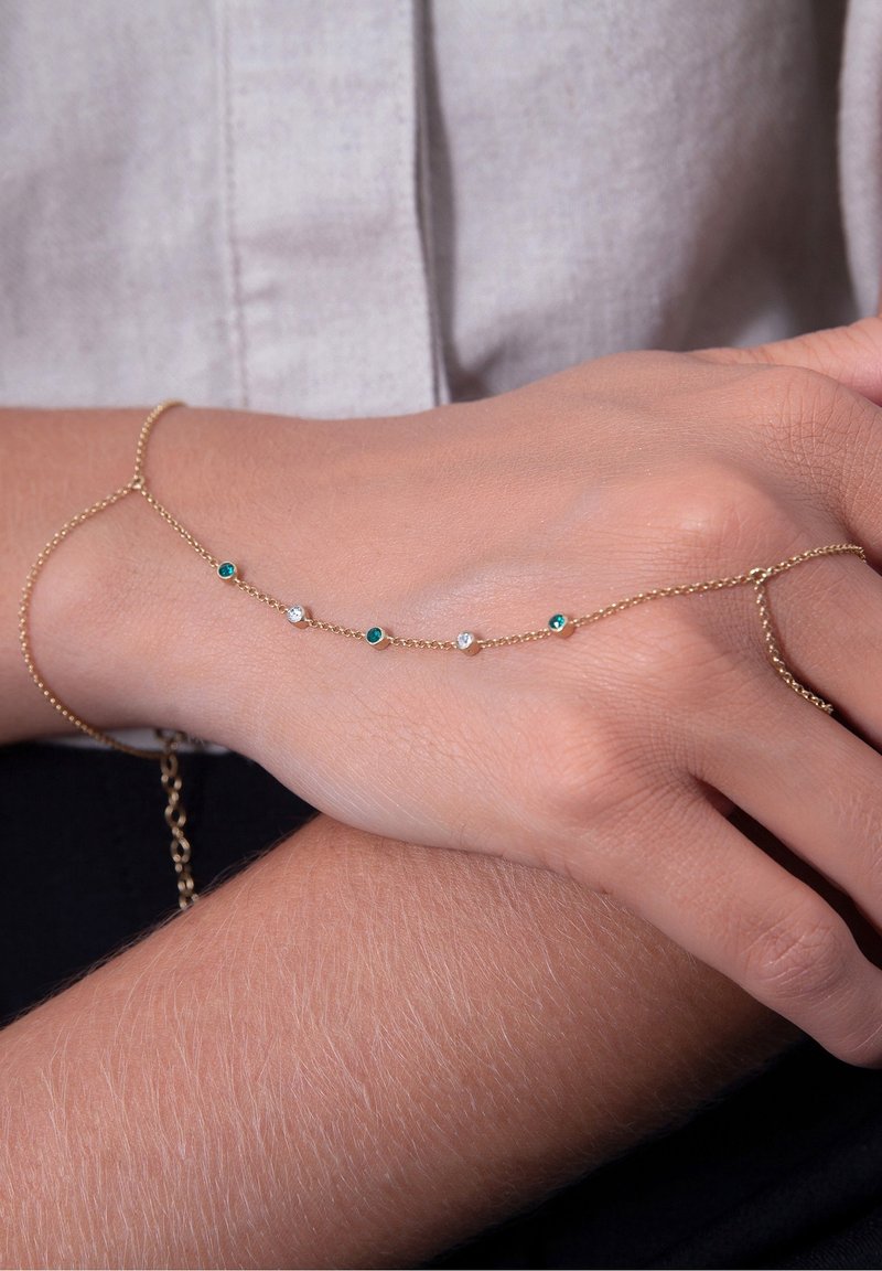 Bracelet chaîne en or délicat avec des petites pierres précieuses vertes et blanches alternées porté au poignet et à la main d'une personne sur un vêtement en tissu clair.