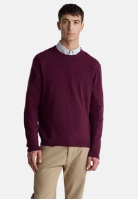 Bordowy sweter wykonany z miękkiej tkaniny, z okrągłym dekoltem i prążkowanym ściągaczem. Model nosi go na koszuli z kołnierzykiem, w połączeniu z beżowymi spodniami.