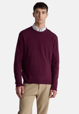 Vínový svetr z měkké látky s kulatým výstřihem a žebrovaným lemem. Model ho nosí přes košili s límečkem, v kombinaci s béžovými kalhotami.