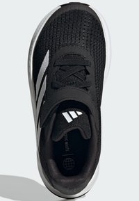 Zapatilla deportiva negra con un upper texturizado, que presenta una correa prominente, acentos grises y una suela blanca. Incluye "Residuos Plásticos" en el interior.