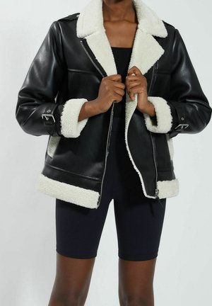 Personne portant une veste en cuir synthétique noire avec bordure en laine blanche, tenant le col de la veste, vêtue d’un short noir ajusté et d’un haut.