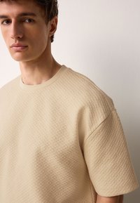 Beige textured t-shirt with a crew neck, featuring a geometric pattern and short sleeves, made from a soft fabric.