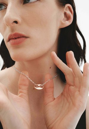 Collar de cadena de plata con un pendant ovalado pulido de diseño entrelazado, usado en un cuello desnudo, con manos que lo sostienen suavemente.