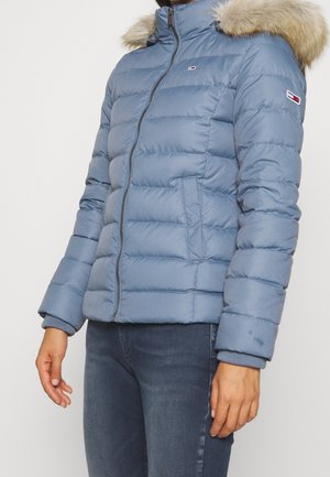 Femme portant une veste matelassée bleu clair avec capuche doublée de fourrure, fermeture éclair, et logos brodés sur la poitrine et la manche, associée à un jean bleu.