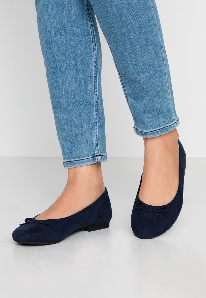 Pieds portant des ballerines bleu marine avec de petits nœuds, assorties à un jean bleu clair retroussé sur un fond de sol uni.