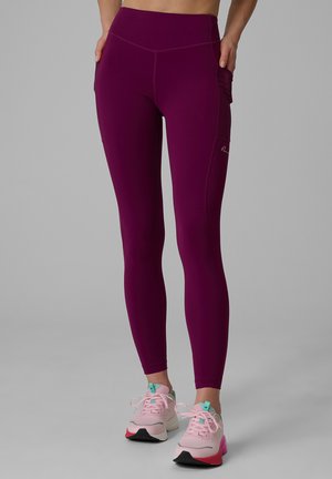 Femme portant un legging violet taille haute avec des poches latérales et des chaussures de sport rose clair, debout sur une surface grise.